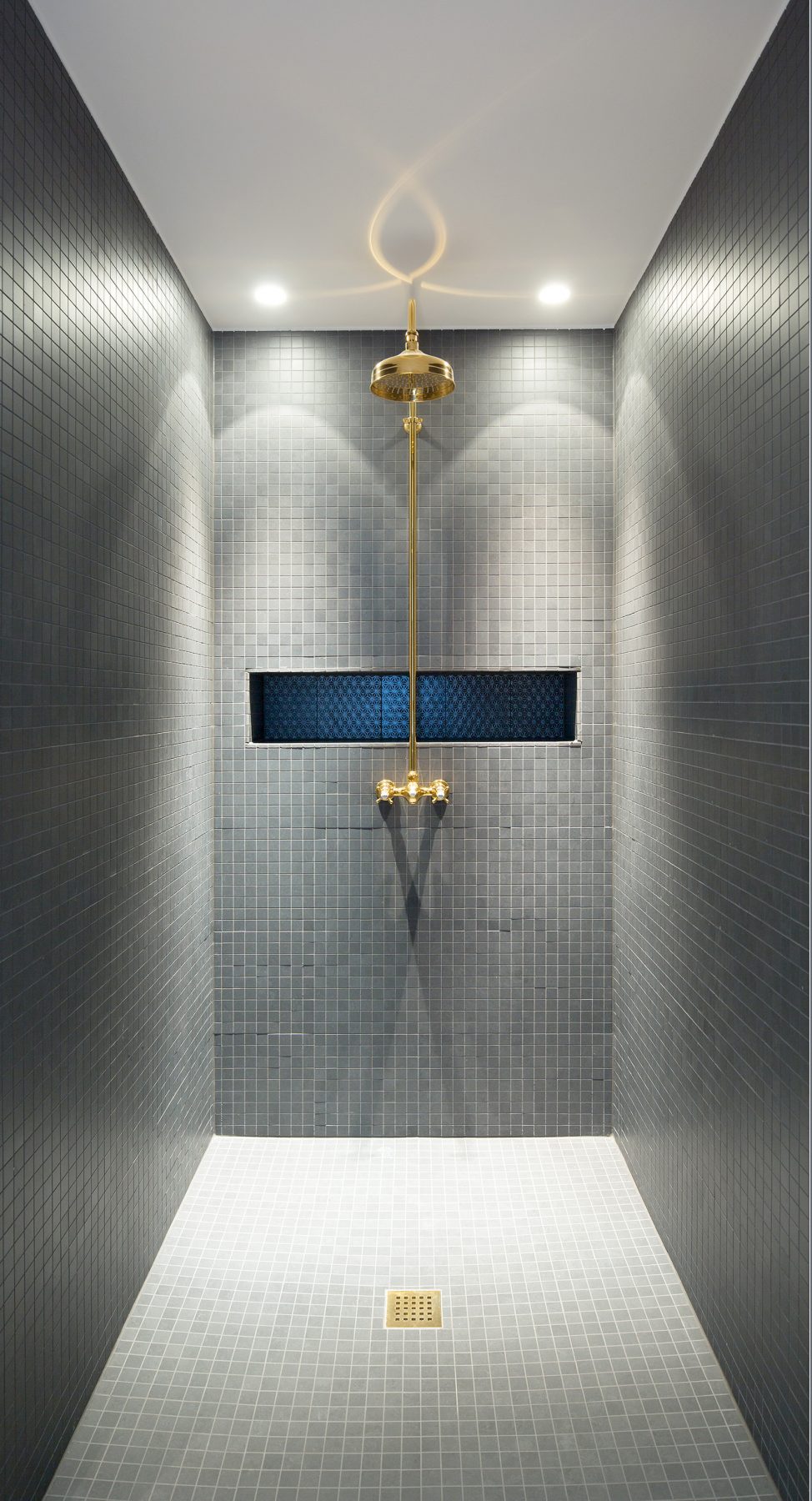 Sprchový kút je obložený malými sivými obkladačkami ktoré nechávajú vyniknúť sprchovú hlavicu batériu a odtokovú mriežku z mosadze.