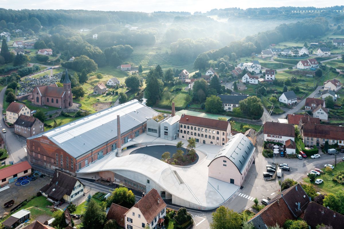 Dvojica bývalých továrenských budov po novom prepája na mieste liaty betónový povrch pripomínajúci vlnu, ktorú možno vnímať aj ako metaforu ťahania rozžeraveného skla.