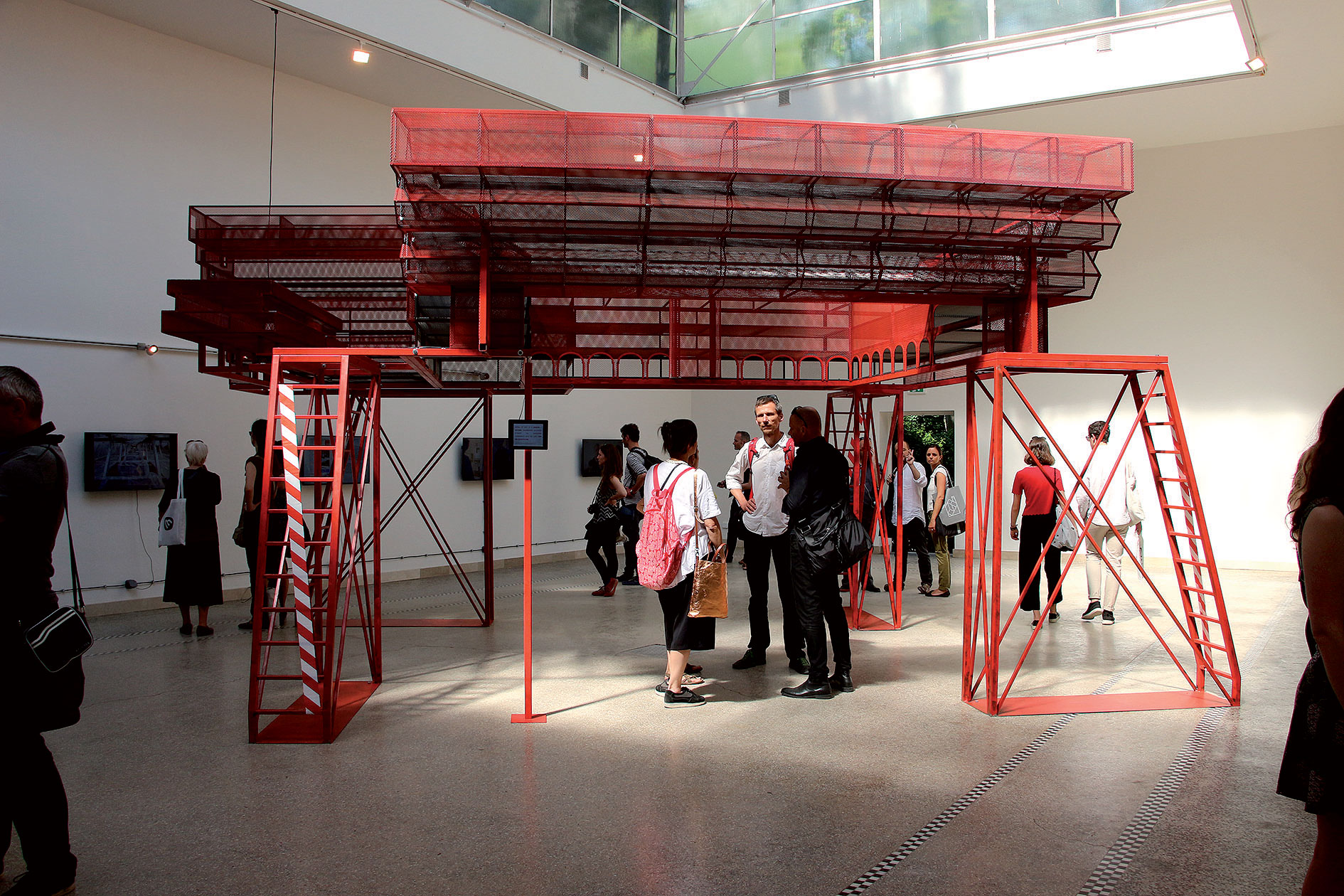Záber počas neformálneho otvorenia pavilónu ČR a SR na benátskom bienále. Ústredným objektom je trojrozmerný model SNG v červenej farbe, ktorý v zmenšenej mierke zodpovedá postavenému areálu.